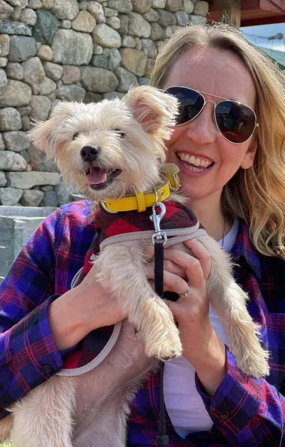 A smiling team member in a plaid shirt holding her small terrier outdoors